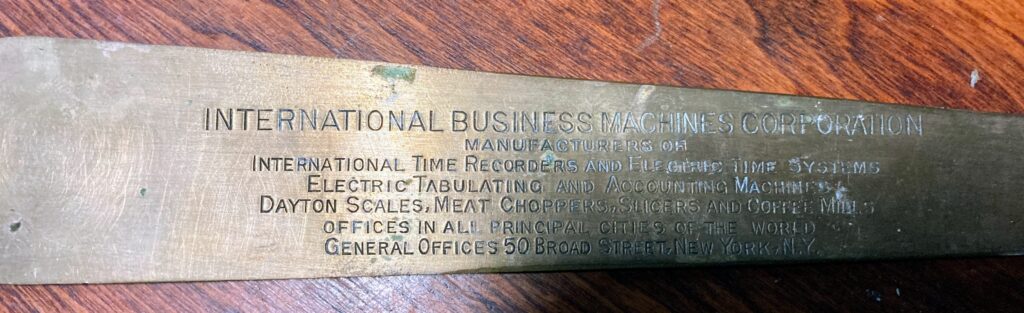 A vintage brass IBM nameplate engraved with early product lines, including time recorders, tabulating machines, scales, meat choppers, slicers, and coffee mills, displayed on a wooden surface.